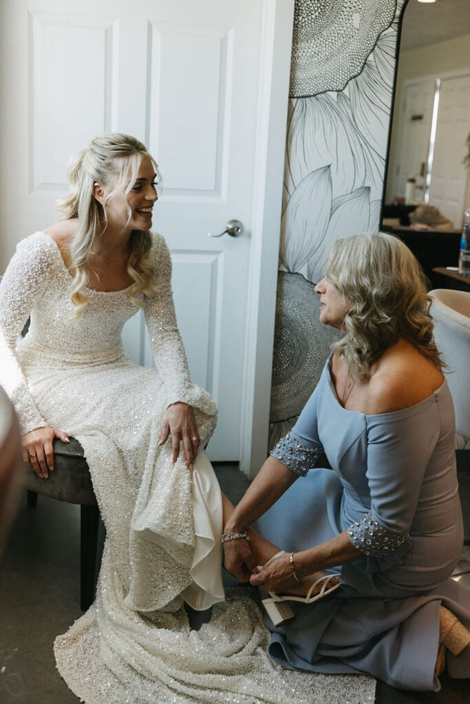 Mother of the bride assists bride in putting shoes on.